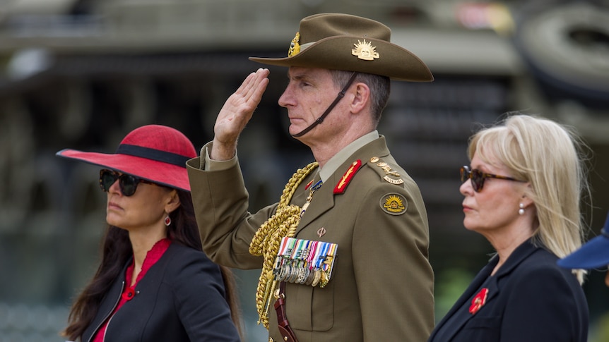 Angus Campbell, in uniform and standing in the middle of two women, saluting. Photo is taken from the left hand side