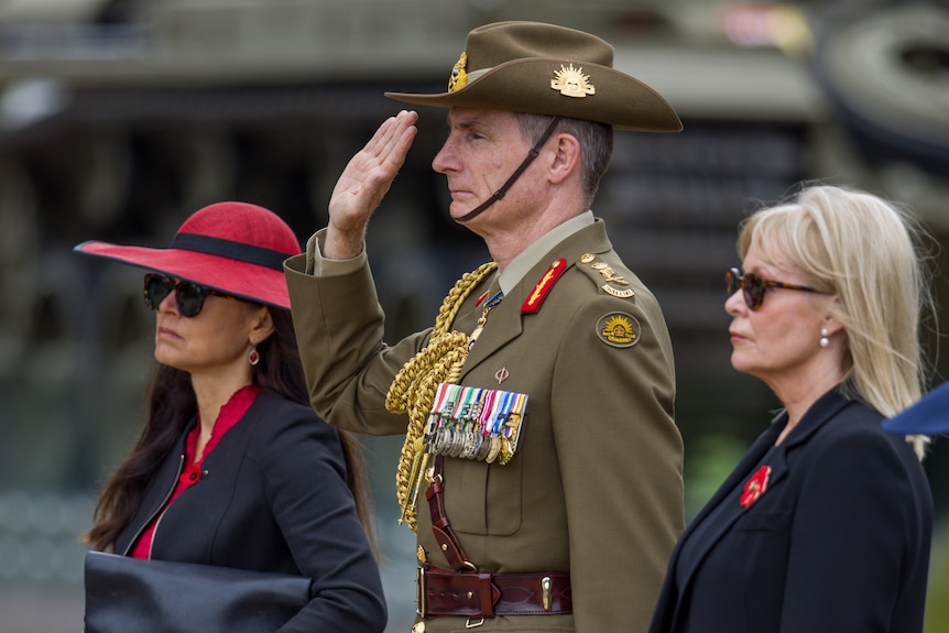 Angus Campbell, in uniform and standing in the middle of two women, saluting. Photo is taken from the left hand side