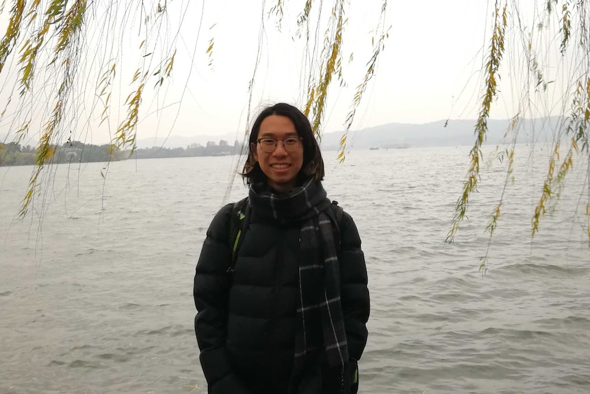 A Chinese man stands in front of a lake and beneath the leaves on a tree.