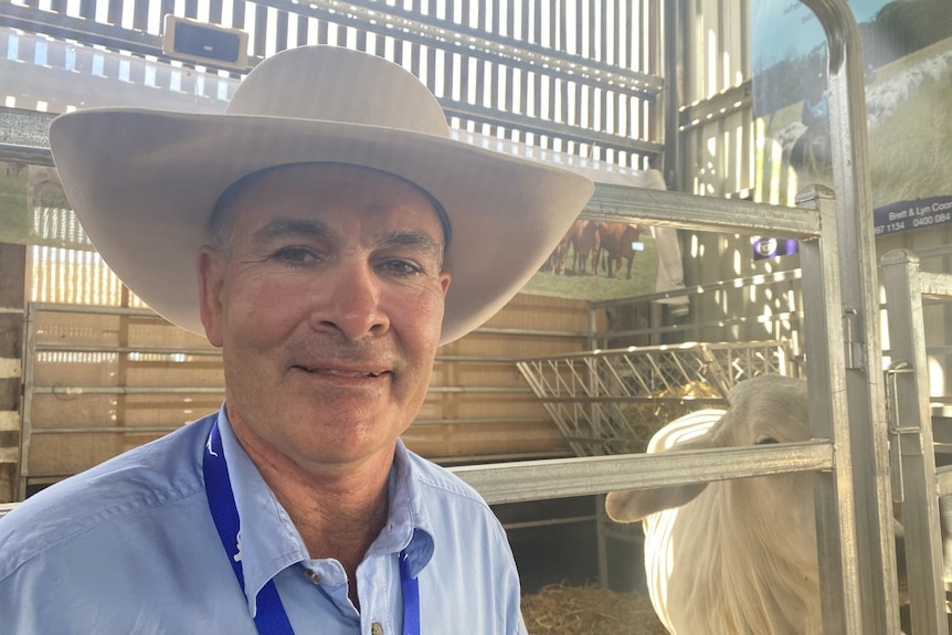 A man smiles. He is wearing a cowboy hat. There is pens and a cow in the background.