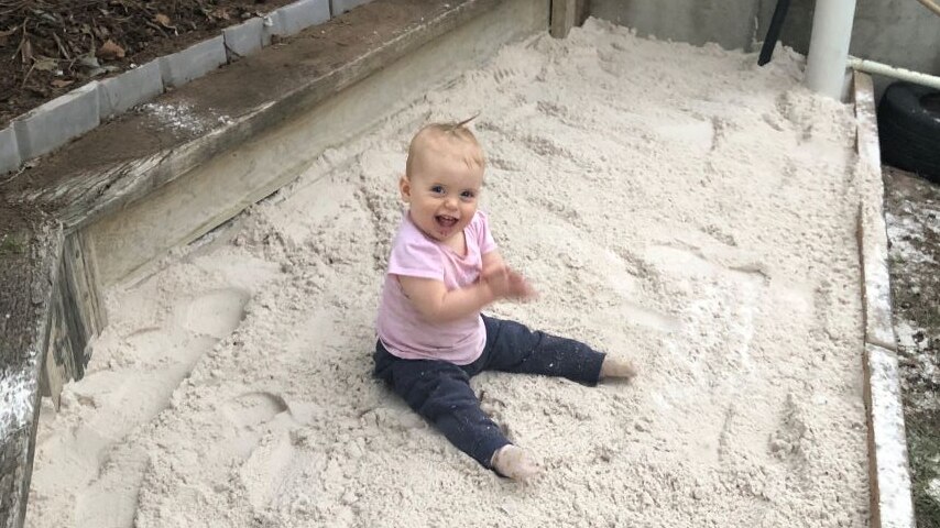 A one year old child sits in a new home made sandpit clapping and smiling