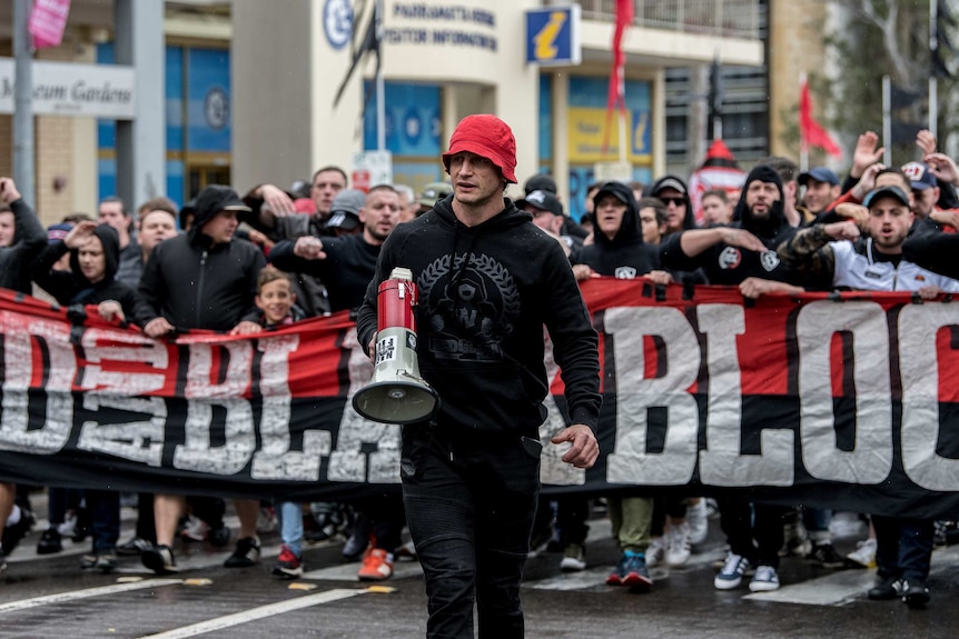 A man with a red hat holds a megaphone and stands in front of the RBB parade.