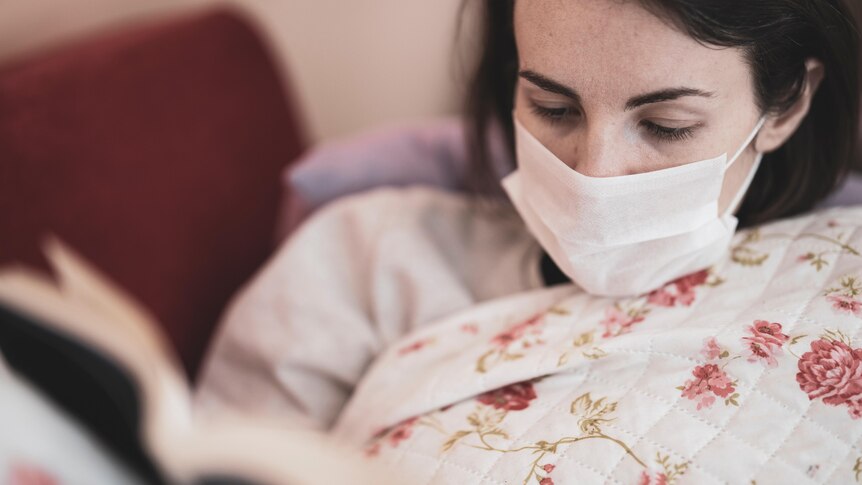 A woman is reading a book and has a face mask on