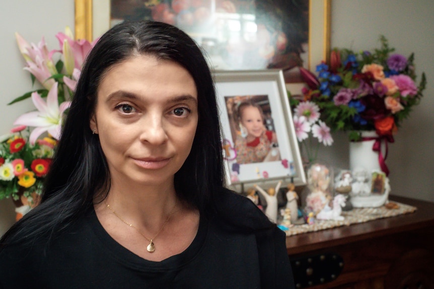 A woman sits in front of flowers and a photo of a young girl. 