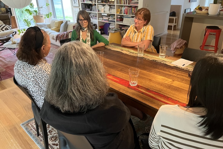 Five women sit around a table talking, only Mara and Litsa are identifiable.