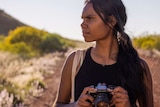 An Aboriginal woman holding a camera, looking away, standing outside, scenery behind her