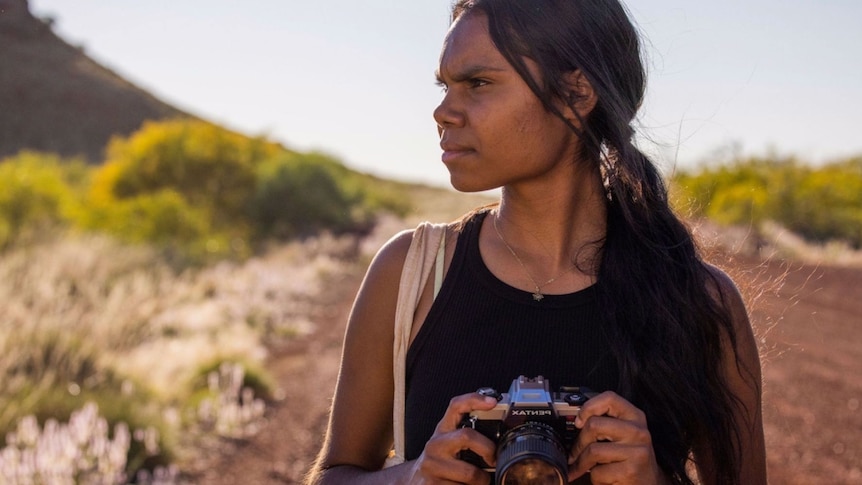An Aboriginal woman holding a camera, looking away, standing outside, dry, dusty scenery behind her.