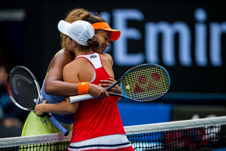 Two tennis players meet, holding their racquets while leaning over the net to hug each other after a match.