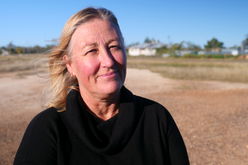 A woman with blonde hair stands in the outback, smiling.