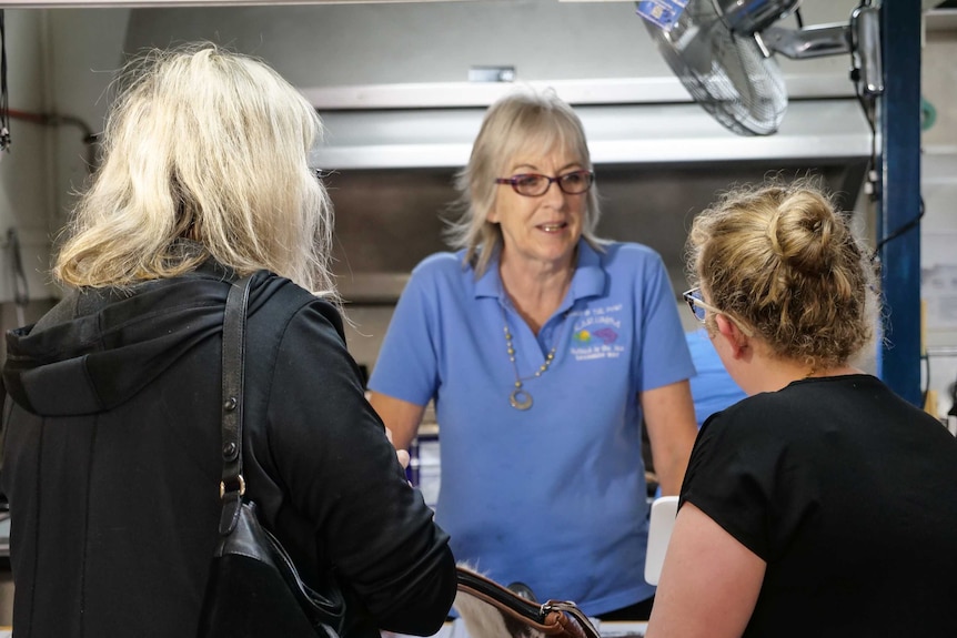 An image of an older woman with blonde hair serving two people, both with blonde hair, at a counter.