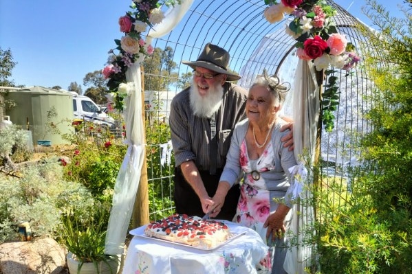 A smiling older couple cut a wedding cake beneath a trellis.