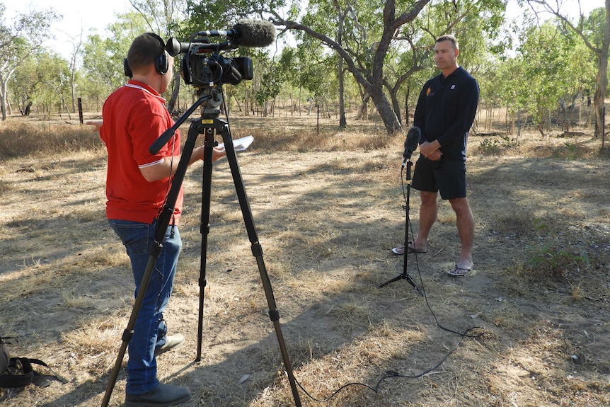 Man in red shirt behind camera talks to another man standing in front of microphone