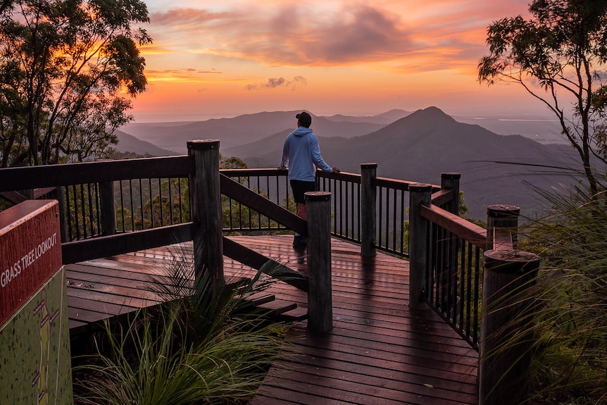 Vibrant sun in the sky, colours reflecing on the board walk, person standing and looking out towards the mountains.