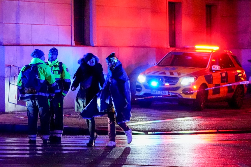 People, one wrapped in a thermal blanket, walk near a university building next to police and a police car. 