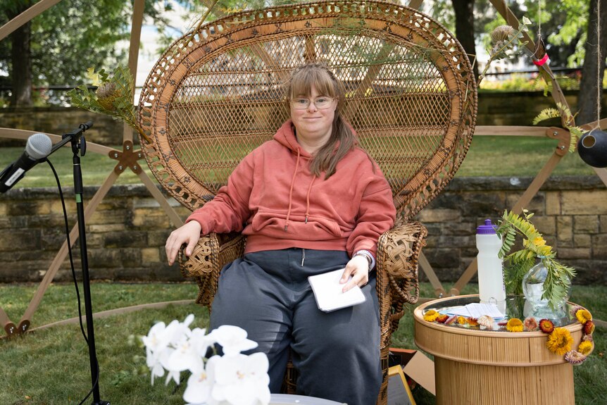 A woman in a salmon jumper sits in a large cane chair 