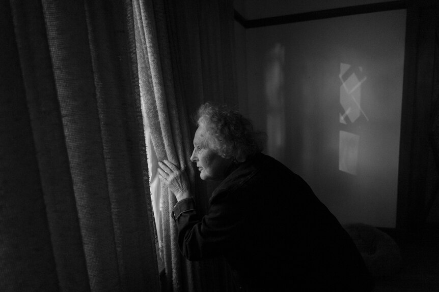Elderly woman looks out a window in a lounge room.