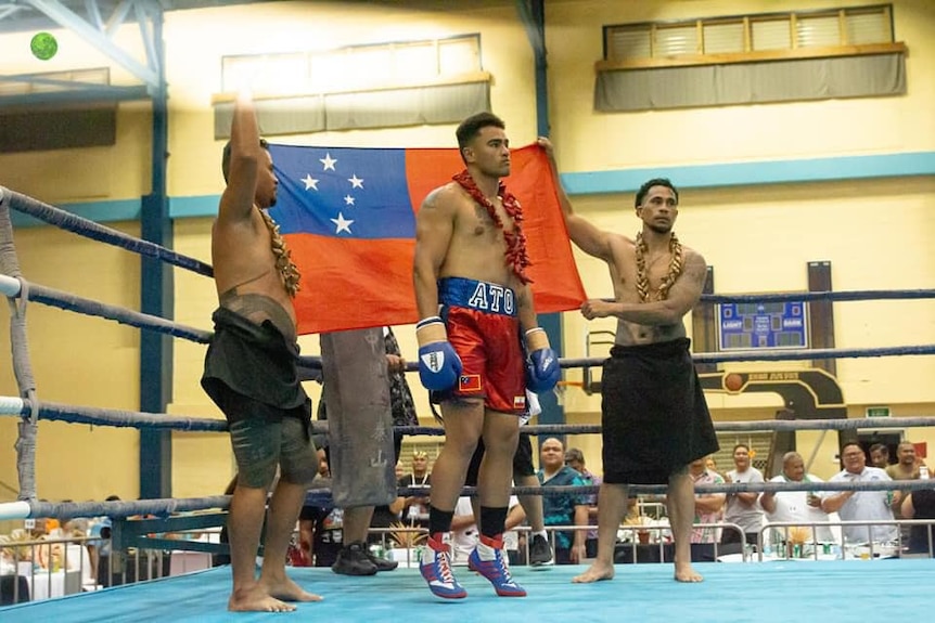 Boxer in red and blue shorts with name Ato stands infront of Samoa flag held by two men in boxing ring. 