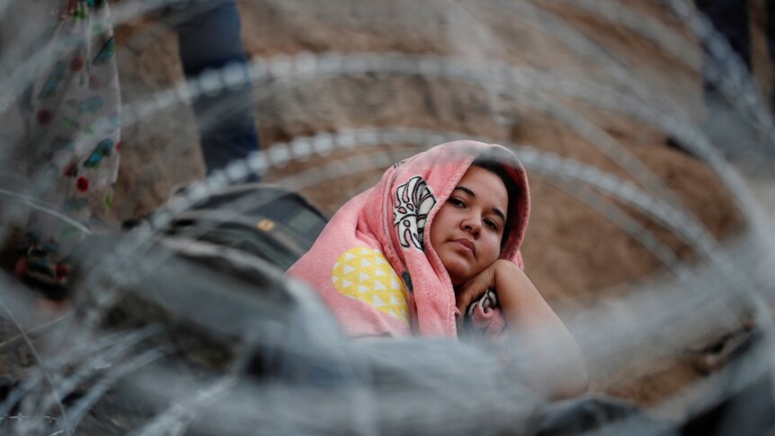 A woman, covering her head in a blanket, sits looking upward, surrounded by barbed wire
