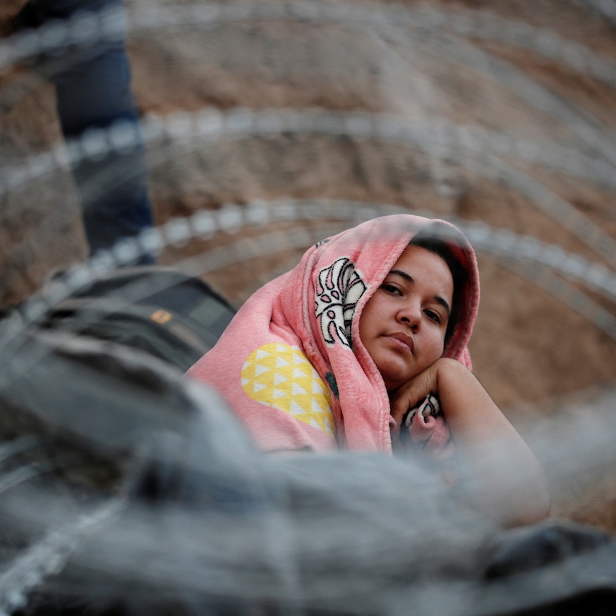 A woman, covering her head in a blanket, sits looking upward, surrounded by barbed wire