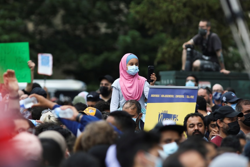 A young girl wearing a pink hijab and a mask holds a mobile phone as people attend during a vigil at the London Mosque.