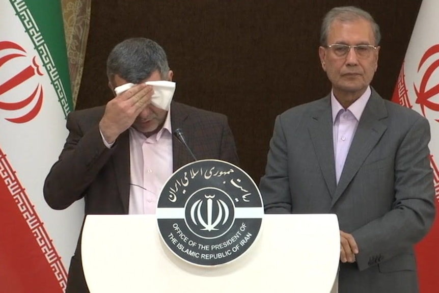 Iraj Harirchi, standing near a podium, holds a cloth to his forehead.