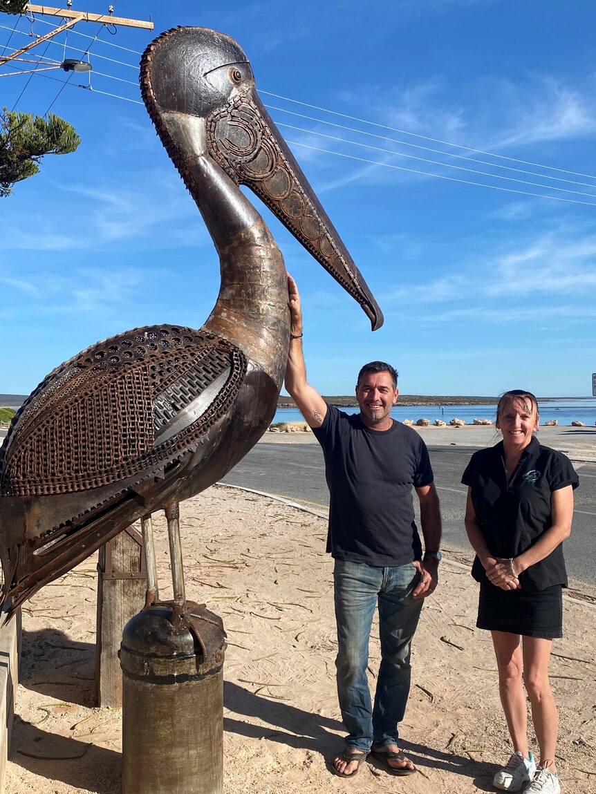 Large bronze pelican on stand with man leaning on it and woman next to him looking at the camera, sunny day, beach background