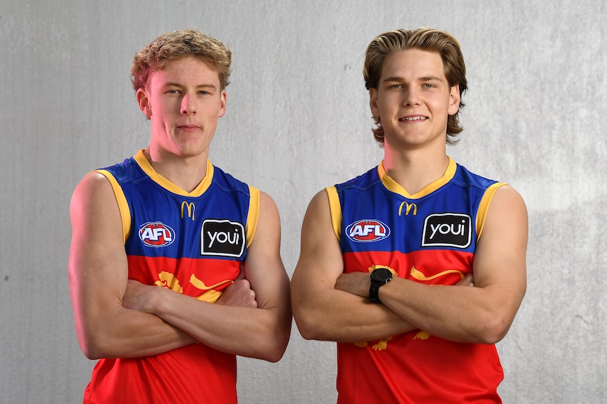 Two young men in Brisbane Lions guernseys stand facing the camera with arms folded.
