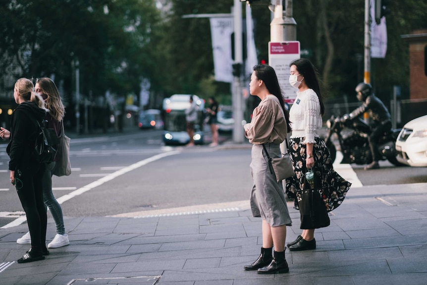 Four women wait to cross the road at traffic lights. One is wearing a face mask and they are standing far apart from each other.
