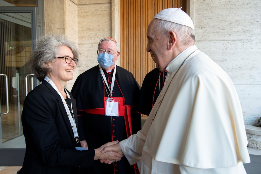 Pope Francis is pictured shaking the hand of a grey-haired woman wearing glasses. Behind them stands a cardinal.