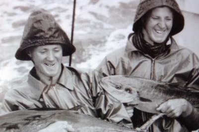 Close up on two young men in wet weather coats and hats on boat at sea holding big fish, ocean in background