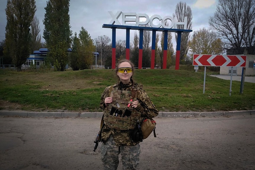 A Ukrainian female soldier poses for a photo against a sign reading Kherson in the background.