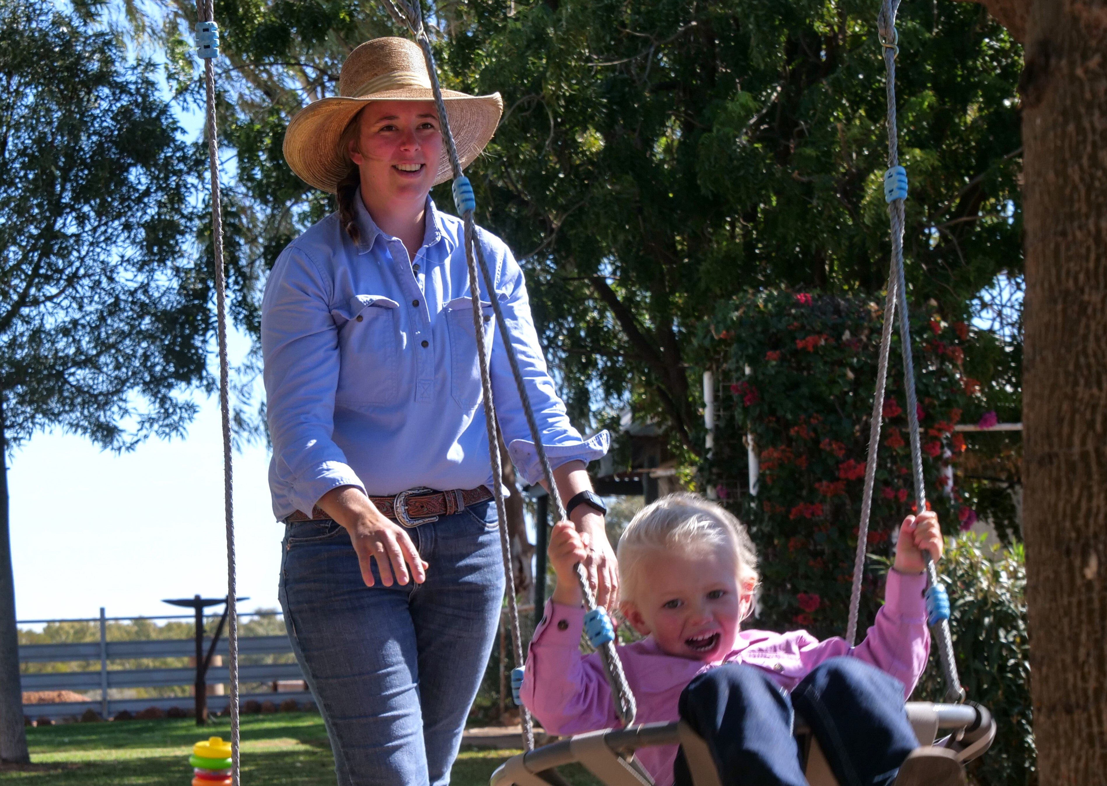 a woman in a blue button up shirt, jeans and cowgirl hat pushes her young blonde haired daughter on a blue swing in a yard