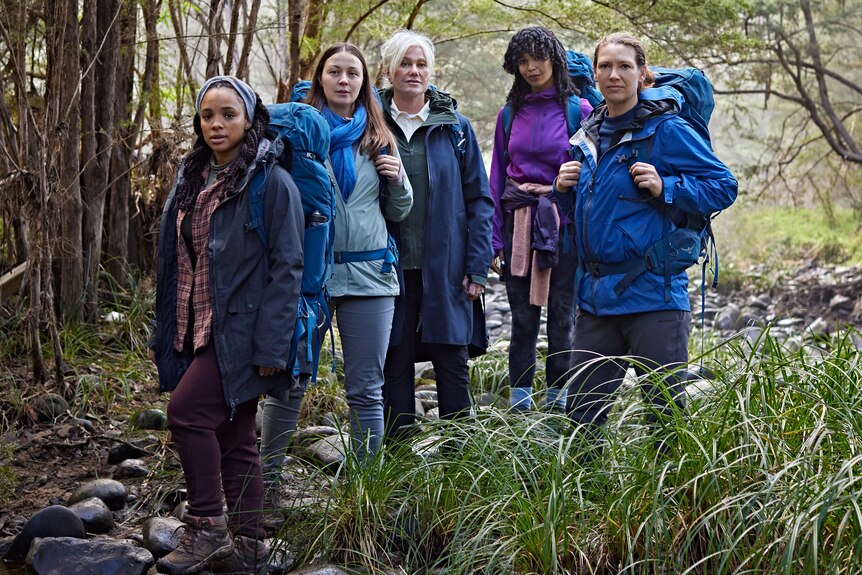 Five women stand in hiking gear in a bushy landscape, staring seriously at the camera