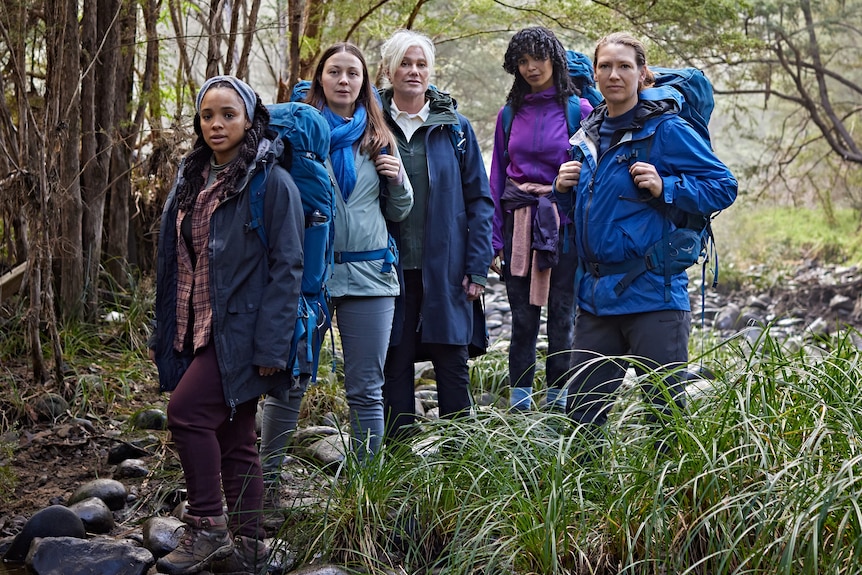 Five women stand in hiking gear in a bushy landscape, staring seriously at the camera