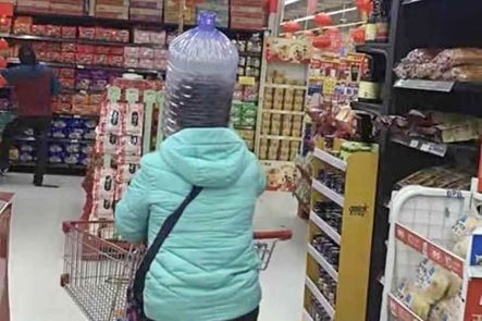 A woman in a Chinese shop walks away from the camera and has a large, empty water bottle covering her head.