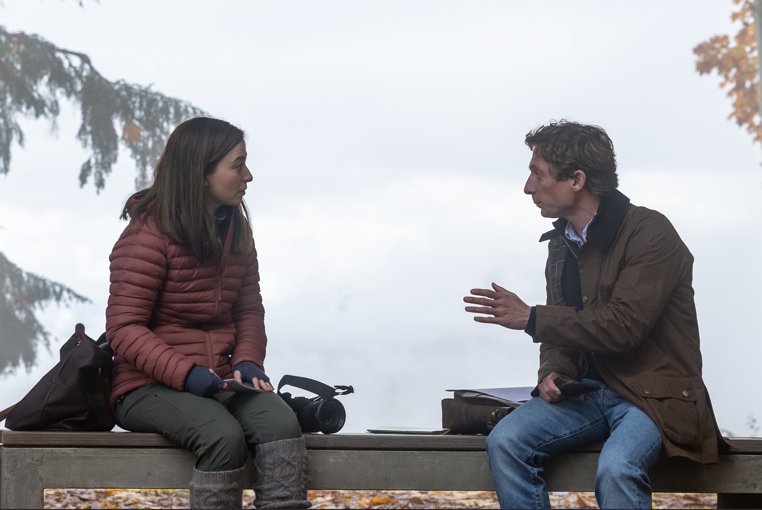 A man and a woman sit facing each other talking on a park bench, wearing wintery clothes.