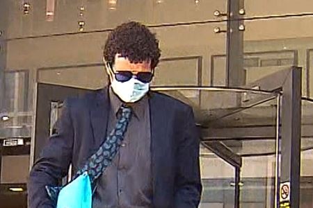 A man with dark, curly hair, wearing a dark suit, sunglasses and a mask, exits a court building.