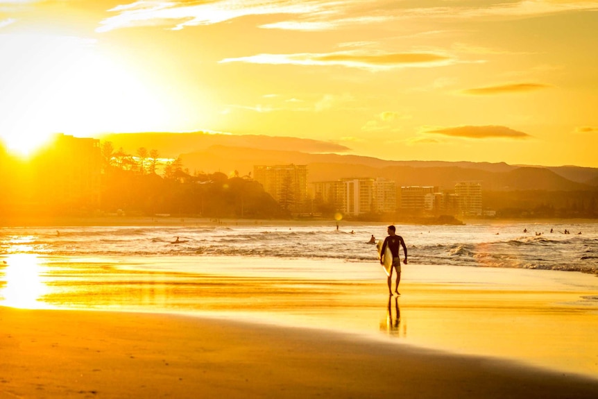 A silhouetted surfer walks along the beach at sunset, the sun reflecting off the shallows.