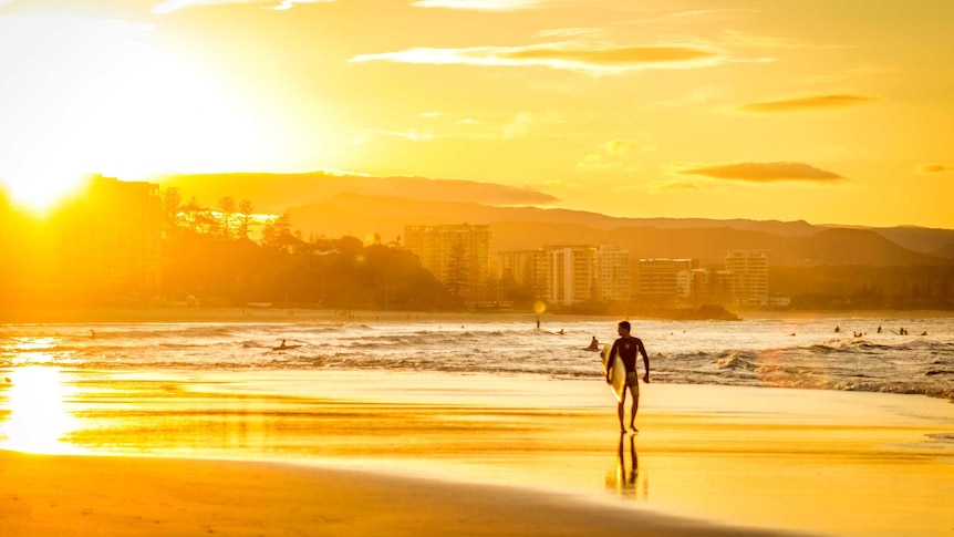 A silhouetted surfer walks along the beach at sunset, the sun reflecting off the shallows.
