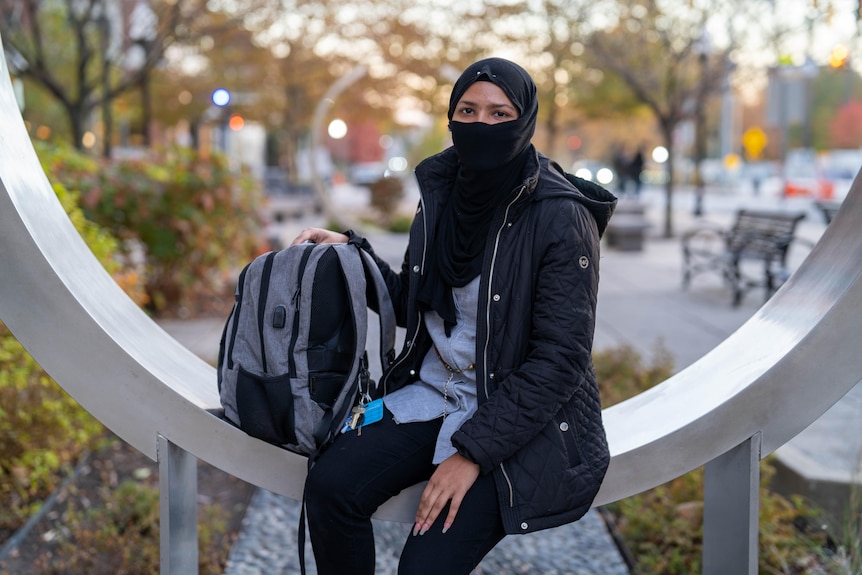Dua, wearing a headscarf and face covering, holds a backpack and sits on a circular metal structure outdoors.