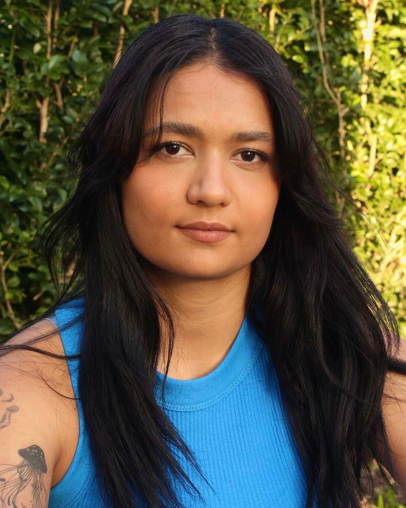 A headshot of a young woman with a blue shirt and long hair with green trees in the background