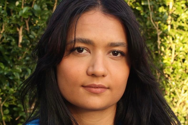 A headshot of a young woman with a blue shirt and long hair with green trees in the background