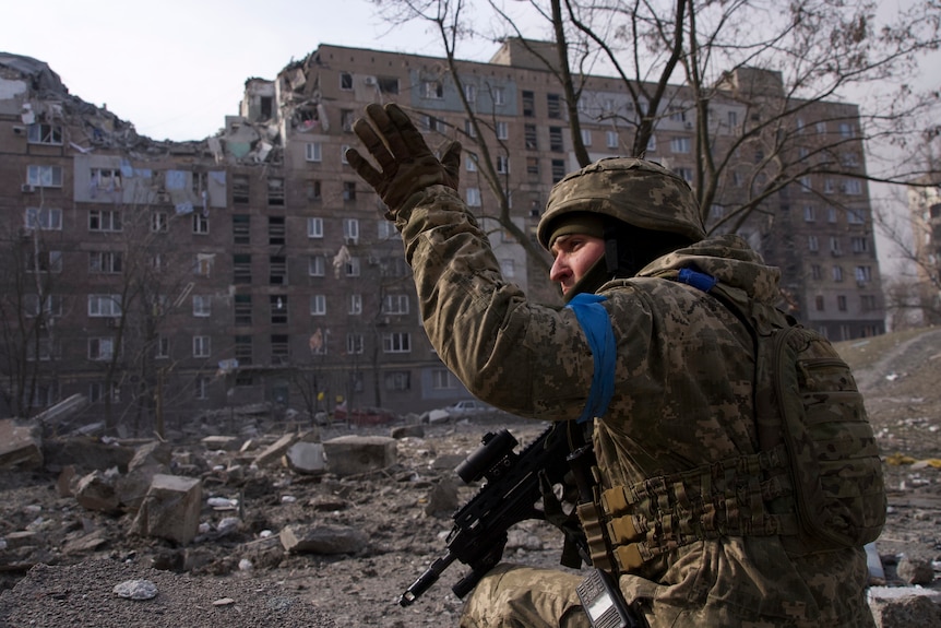 An armed soldier signals with his left hand as he holds a gun in his right hand and looks towards his left.