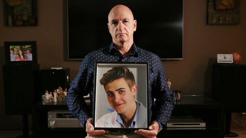 A man sits holding a framed photo of a young man.
