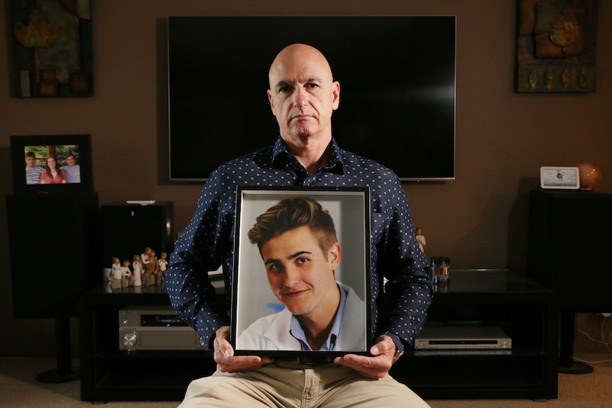 A man sits holding a framed photo of a young man.