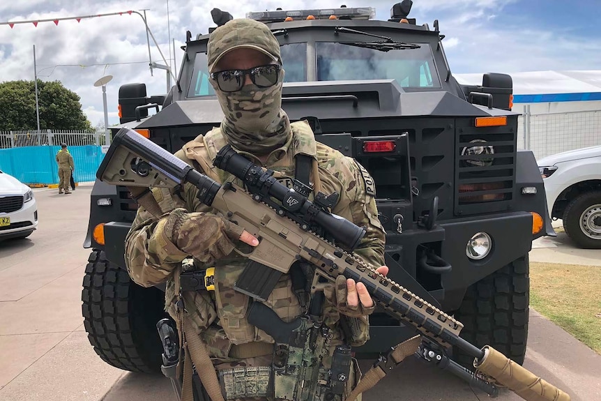 A masked SERT officer holds his rifle to his chest, standing in front of a troop carrier