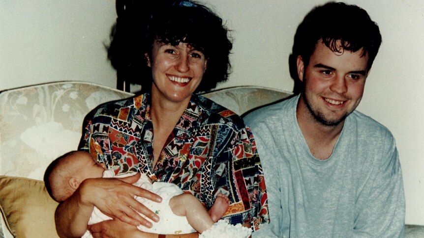 A smiling woman holds a baby in her arms, with a young man smiling sitting next to her.