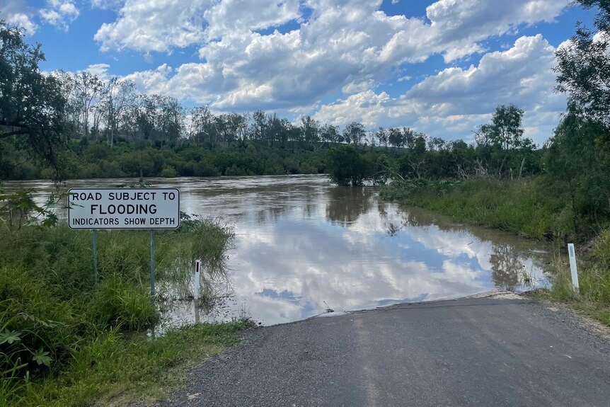 Flood water runs over a road, sign reads "road subject to flooding" 