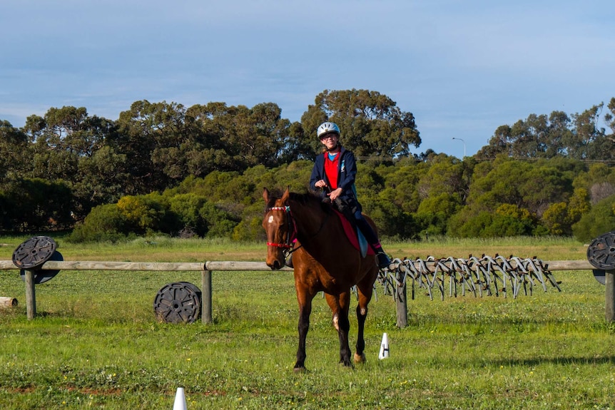 Aleisha sits on top of Ned the horse in a green paddock. She wears a helmet and red shirt.