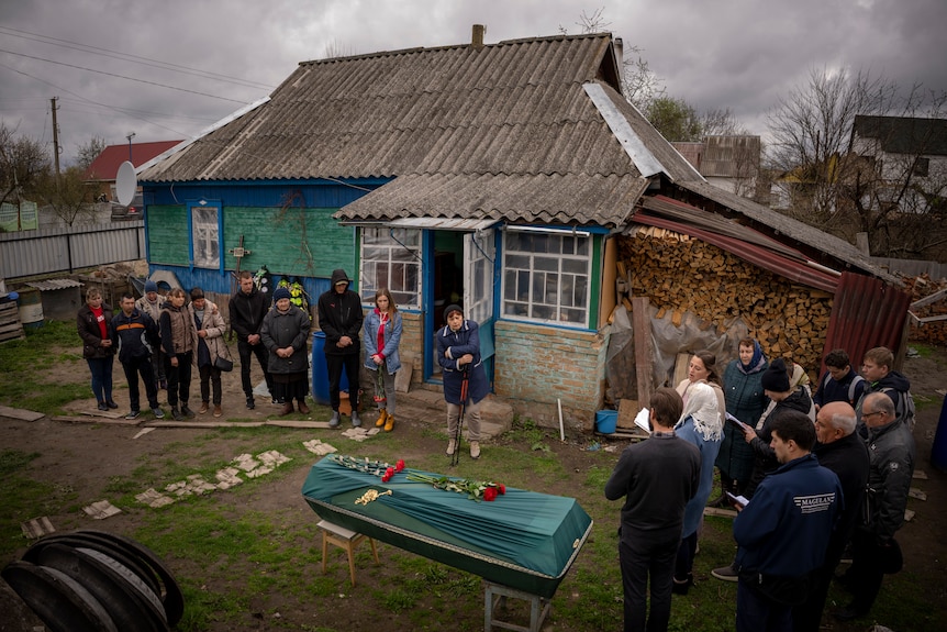 A group of people stand around a flower and silk wrapped coffin in a backyard. 